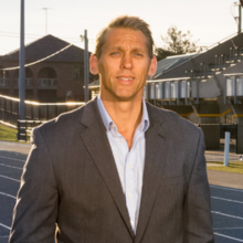 Professor David Lubans - man with a grey suit jacket standing in front of an athletic field with a running track and grandstand