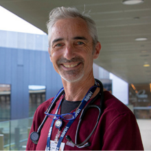 Prof Josh Davis - man with maroon scrubs on and stethoscope around neck smiling at camera