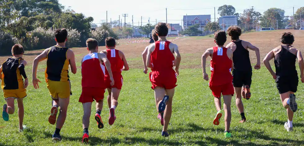 group of teenage boys in sporting clothes and joggers running together. Their backs are turned to us. They are in mid-run at a sporting field