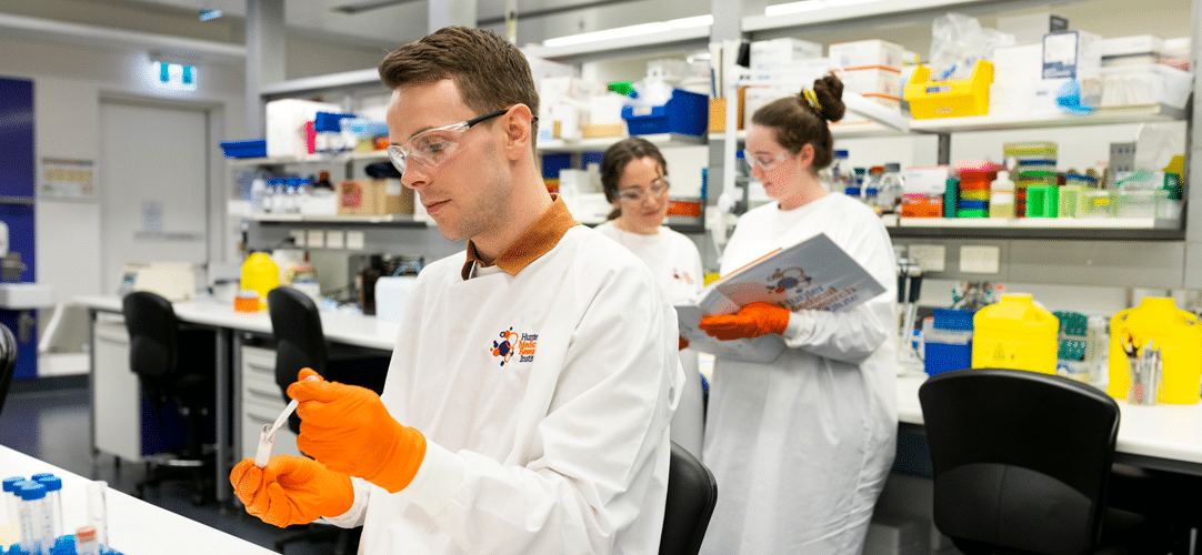 Australia’s medical research institutes deliver billions in economic value and national capability Image features a researcher in a white coat looking at a test tube, with two researchers standing behind them looking at results